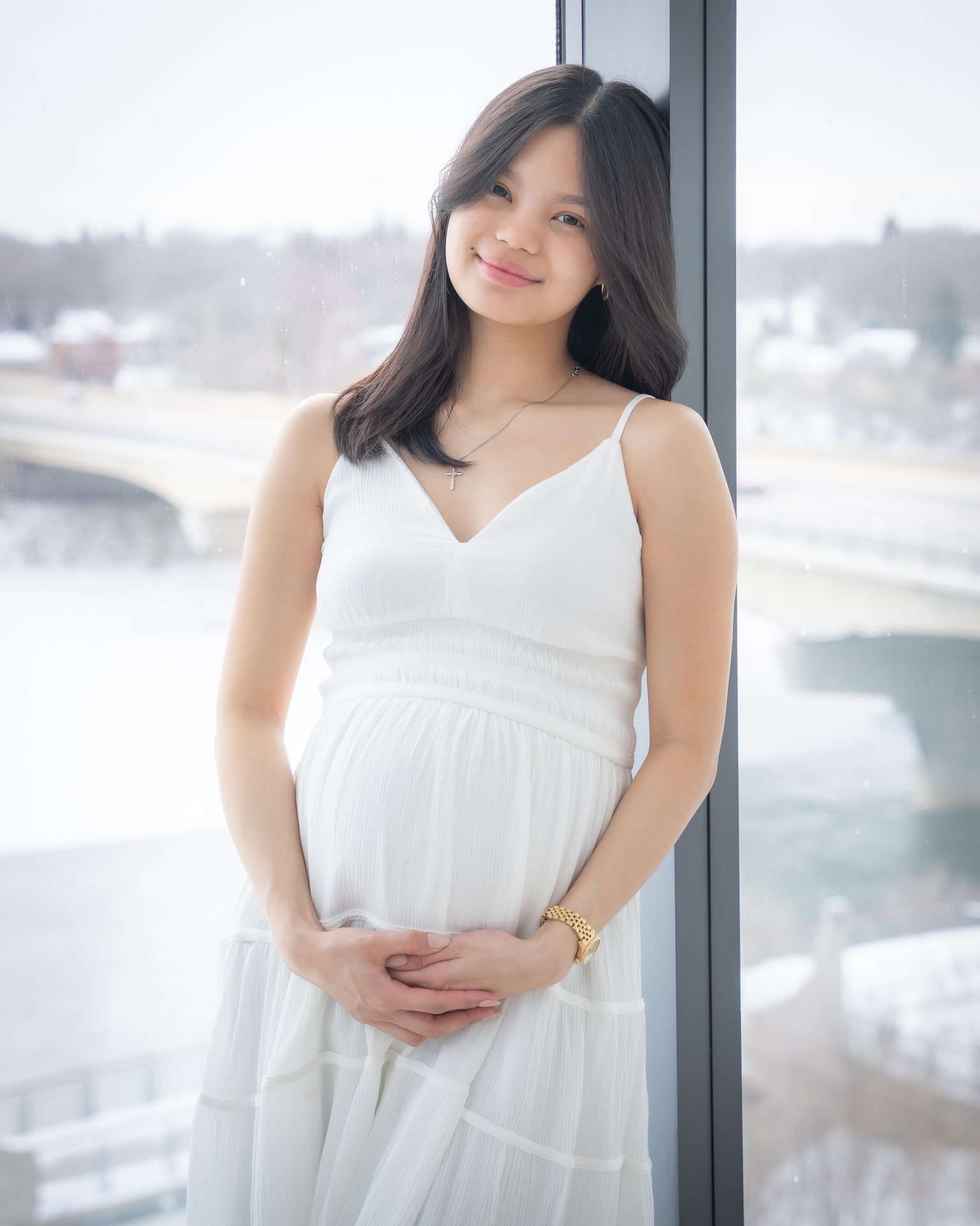 Expecting mother in a white dress by a window overlooking the river in winter