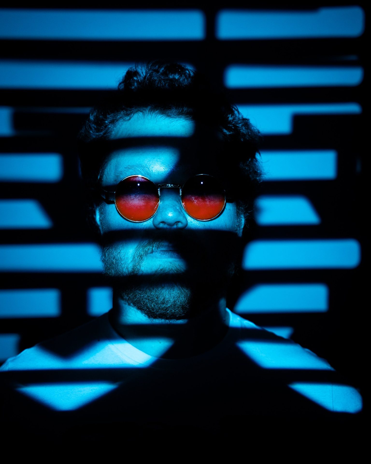 Studio portrait of a man in red round sunglasses, lit in cyan with striped gobo shadows, Saskatoon